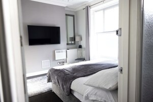 Una habitación de hotel moderna con cama, televisor de pantalla plana, mesita de noche, lámpara y ventana con cortinas.