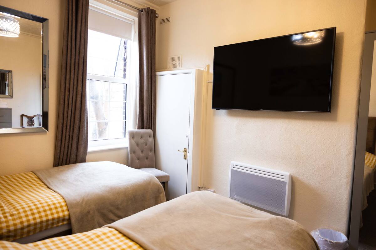 Camera d'albergo con due letti, una televisione, una finestra con tende, un termosifone e uno specchio.