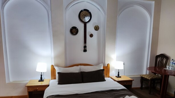 A hotel room with a large bed, two bedside tables with lamps, a wooden nightstand, a chair, and a small table with bottles.