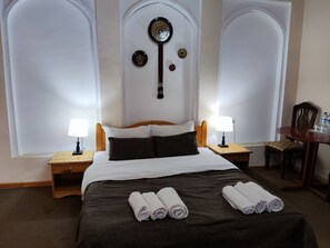 A hotel room with a large bed, two bedside tables with lamps, a wooden nightstand, a chair, and a small table with bottles. - Ziyo-Baxsh Boutique Hotel (Bukhara)