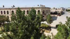 A historic building with arched windows and a flat roof, surrounded by greenery and a paved street.