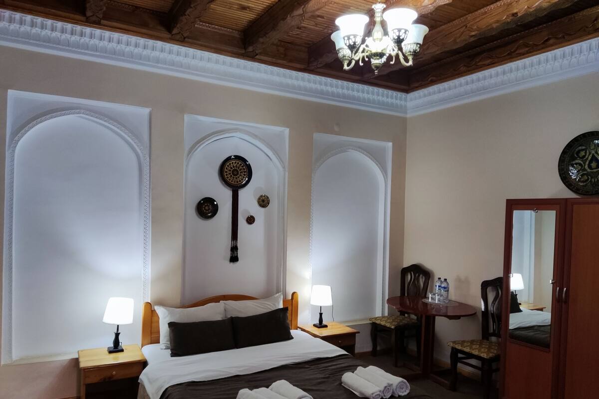 A hotel room with a large bed, two bedside tables with lamps, a wooden wardrobe, and a chandelier.