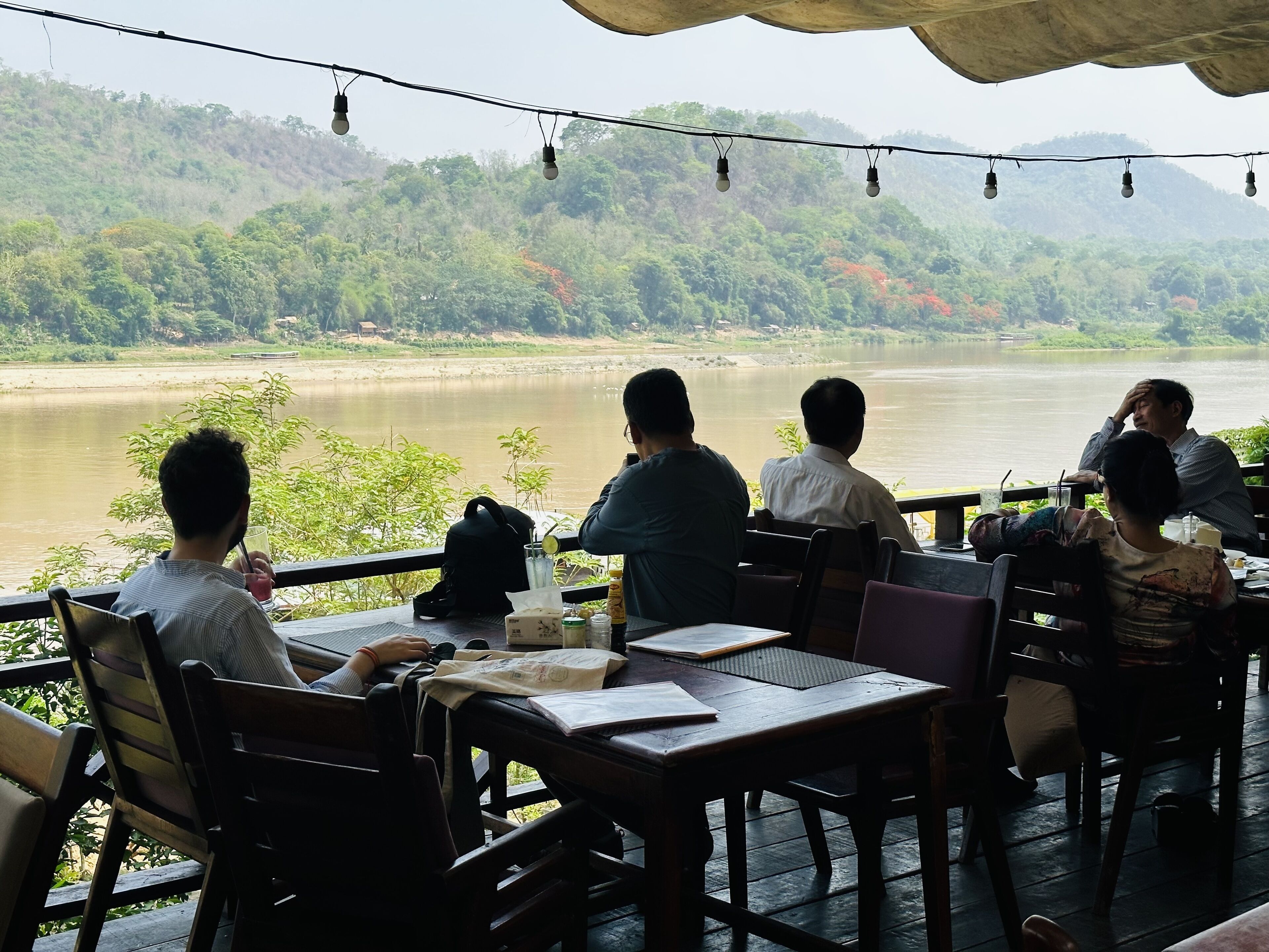 A group of people seated at tables overlooking a river, with a forested hillside in the background.