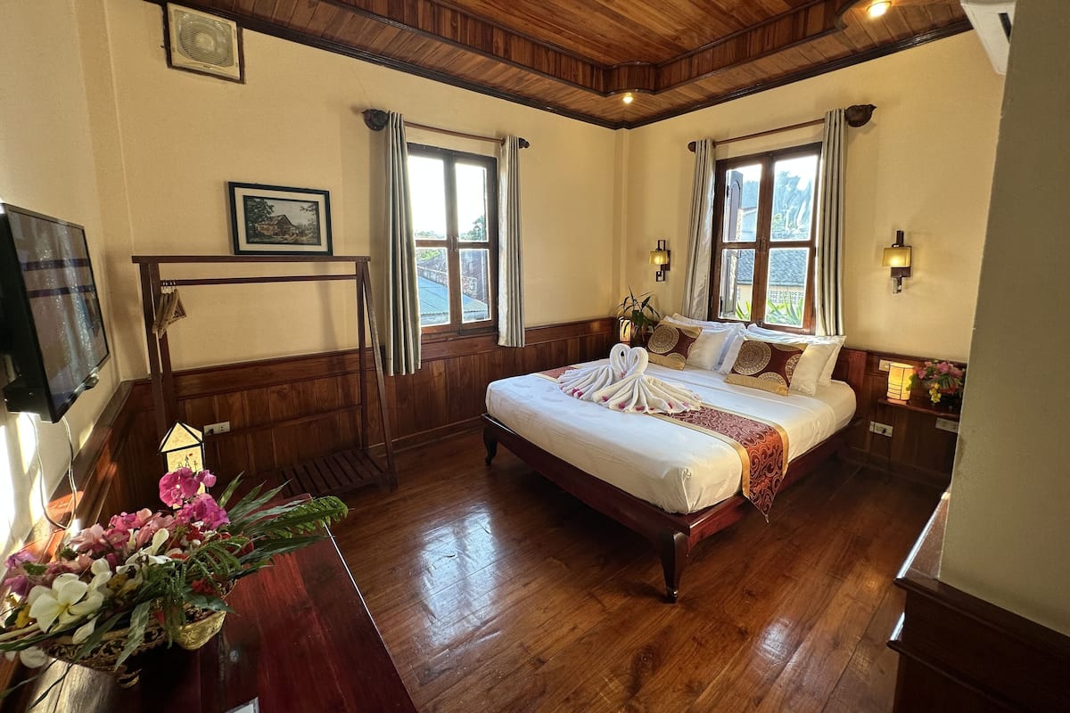 Hotelkamer met een houten plafond, een bed met een witte dekbedovertrek, een houten dressoir, een televisie en een raam met gordijnen.