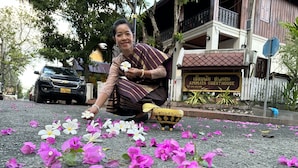 Een vrouw legt bloemen neer op de grond voor een gebouw met een bord waarop staat 'AMMATA GUESTHOUSE'.