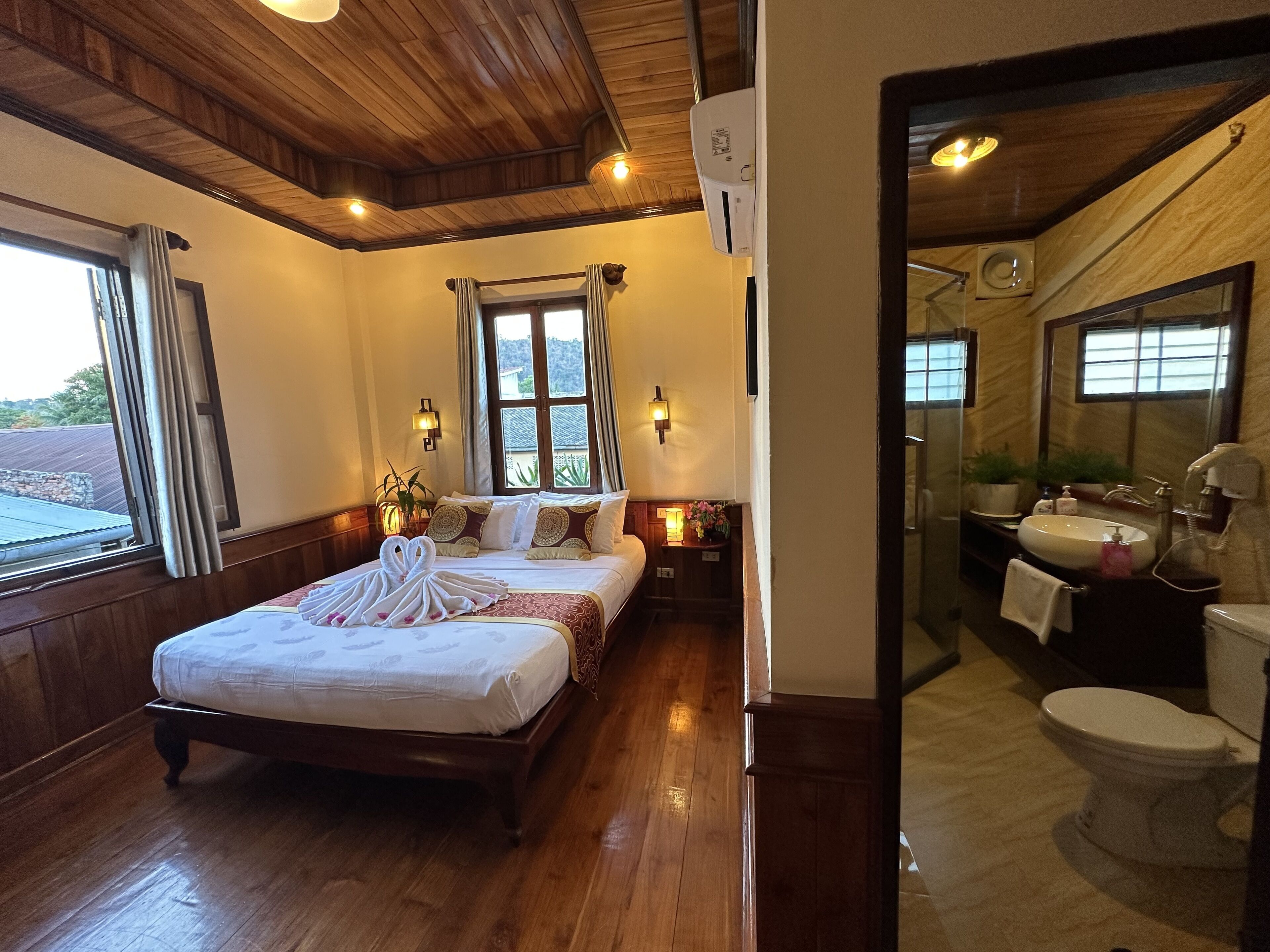 A hotel room with a wooden ceiling, a large bed with a white comforter, two bedside tables with lamps, a window with curtains, and a view of greenery outside.