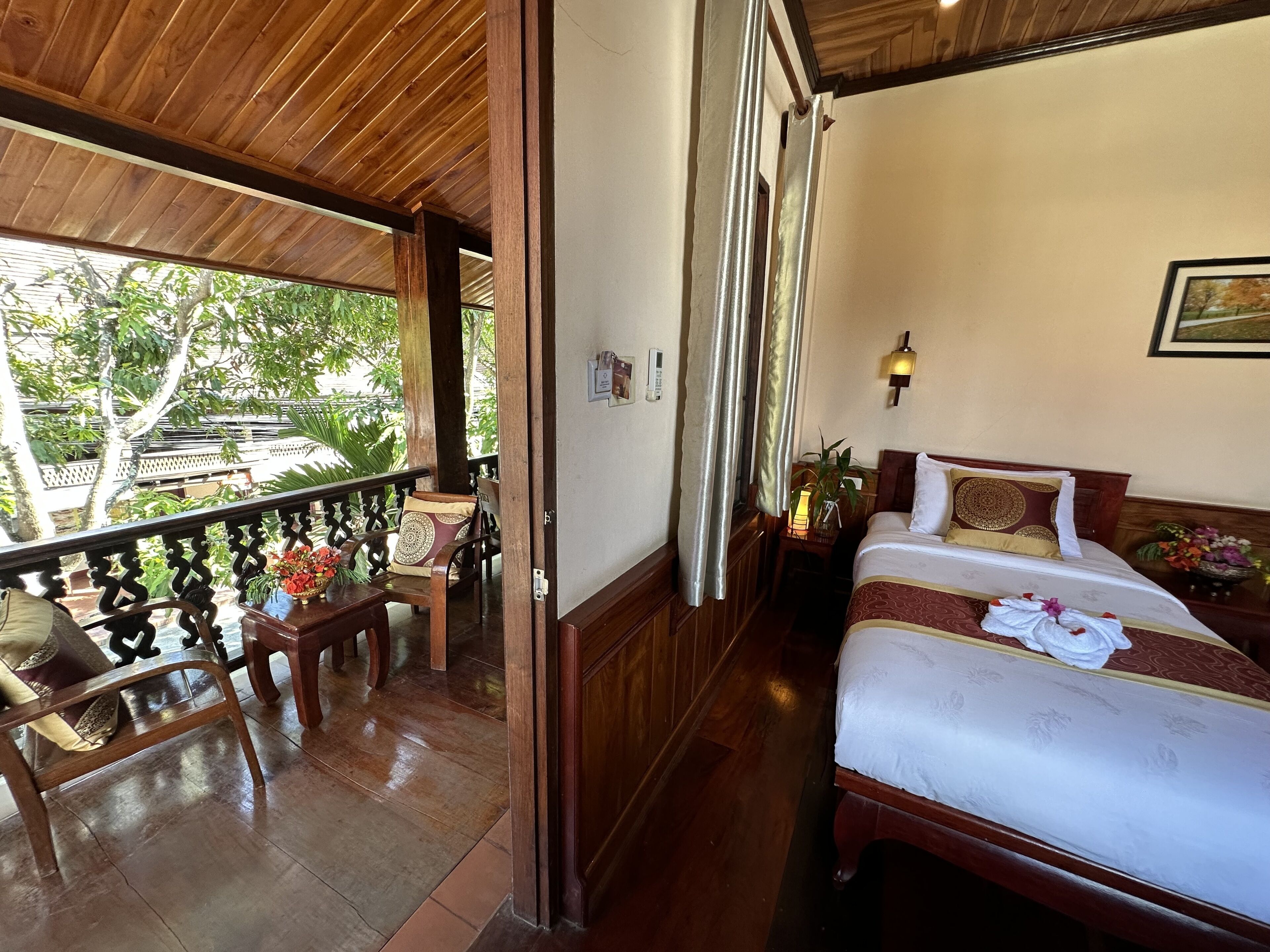 A hotel room with a balcony, wooden furniture, a bed with a white and red bedspread, and a wall-mounted light fixture.