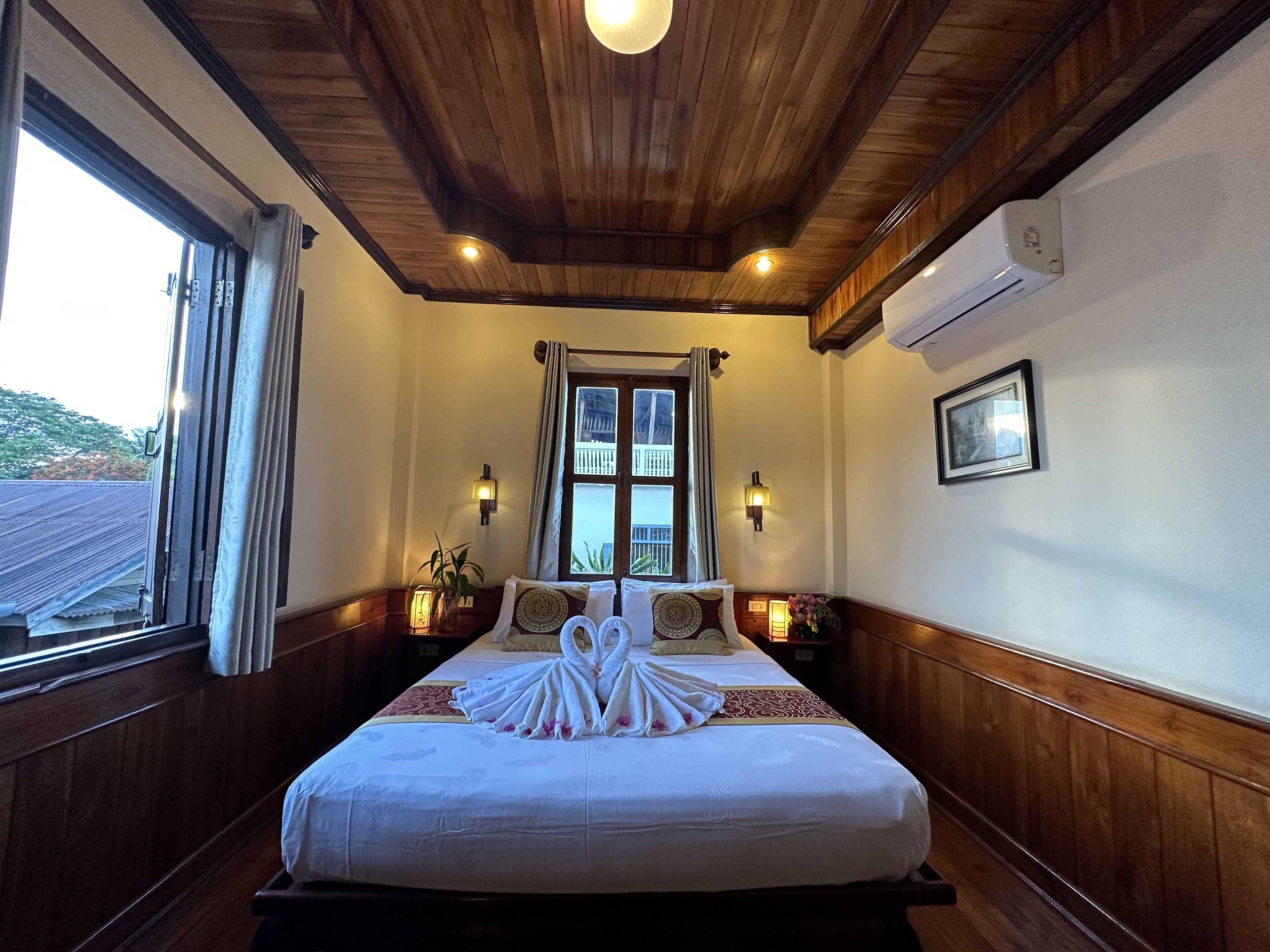 A hotel room with a wooden ceiling, a large bed with white bedding and a towel arrangement, two wall-mounted lamps, a window with curtains, and a framed picture on the wall.