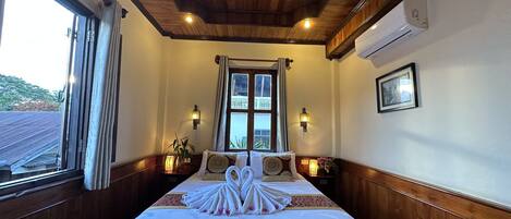 Une chambre d’hôtel avec un plafond en bois, un grand lit avec une literie blanche et des serviettes disposées de manière décorative, deux lampes fixées au mur, une fenêtre avec des rideaux et un tableau encadré accroché au mur.