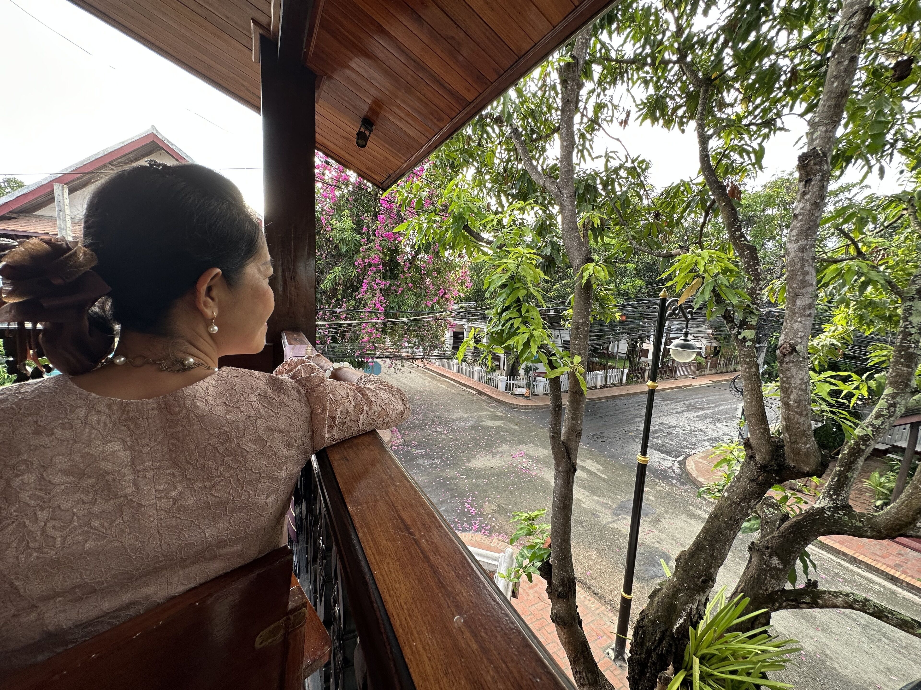 Une personne appuyée sur la balustrade d’un balcon, d’où l’on aperçoit une rue bordée d’arbres et un bâtiment en arrière-plan.
