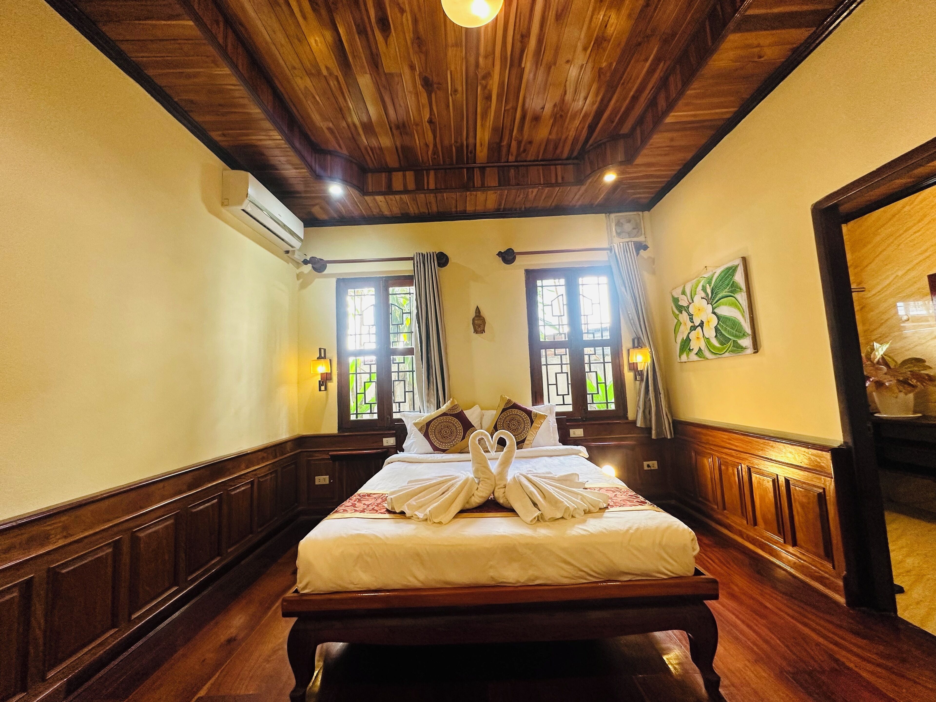 A hotel room with a wooden ceiling, a large bed with white bedding, a bench at the foot of the bed, and a window with curtains.