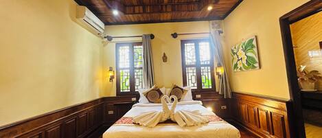 A hotel room with a wooden ceiling, a large bed with white bedding, a bench at the foot of the bed, and a window with curtains.