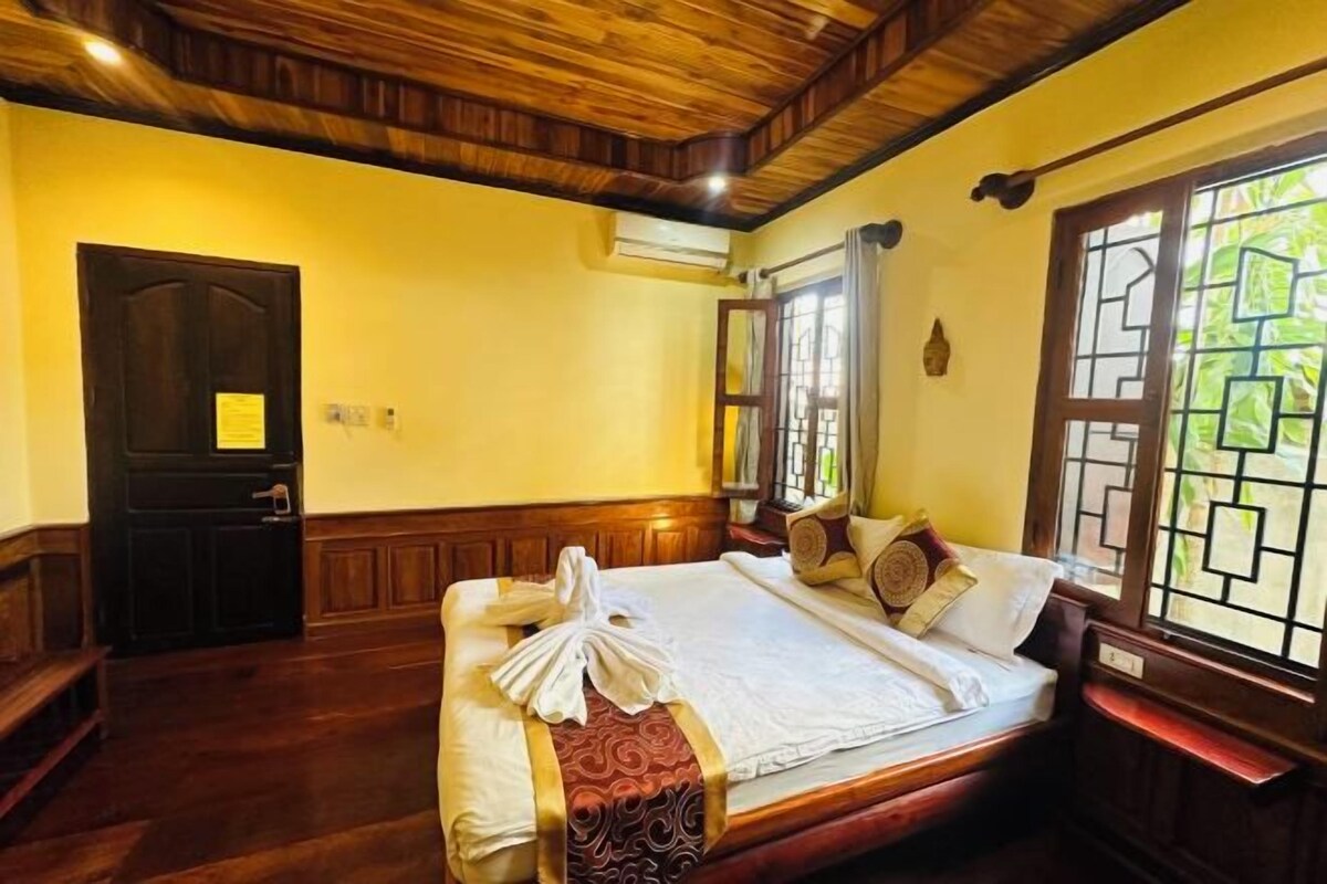A room with a wooden ceiling, a bed with a white and red bedspread, a wooden door, and a window with a view of greenery.