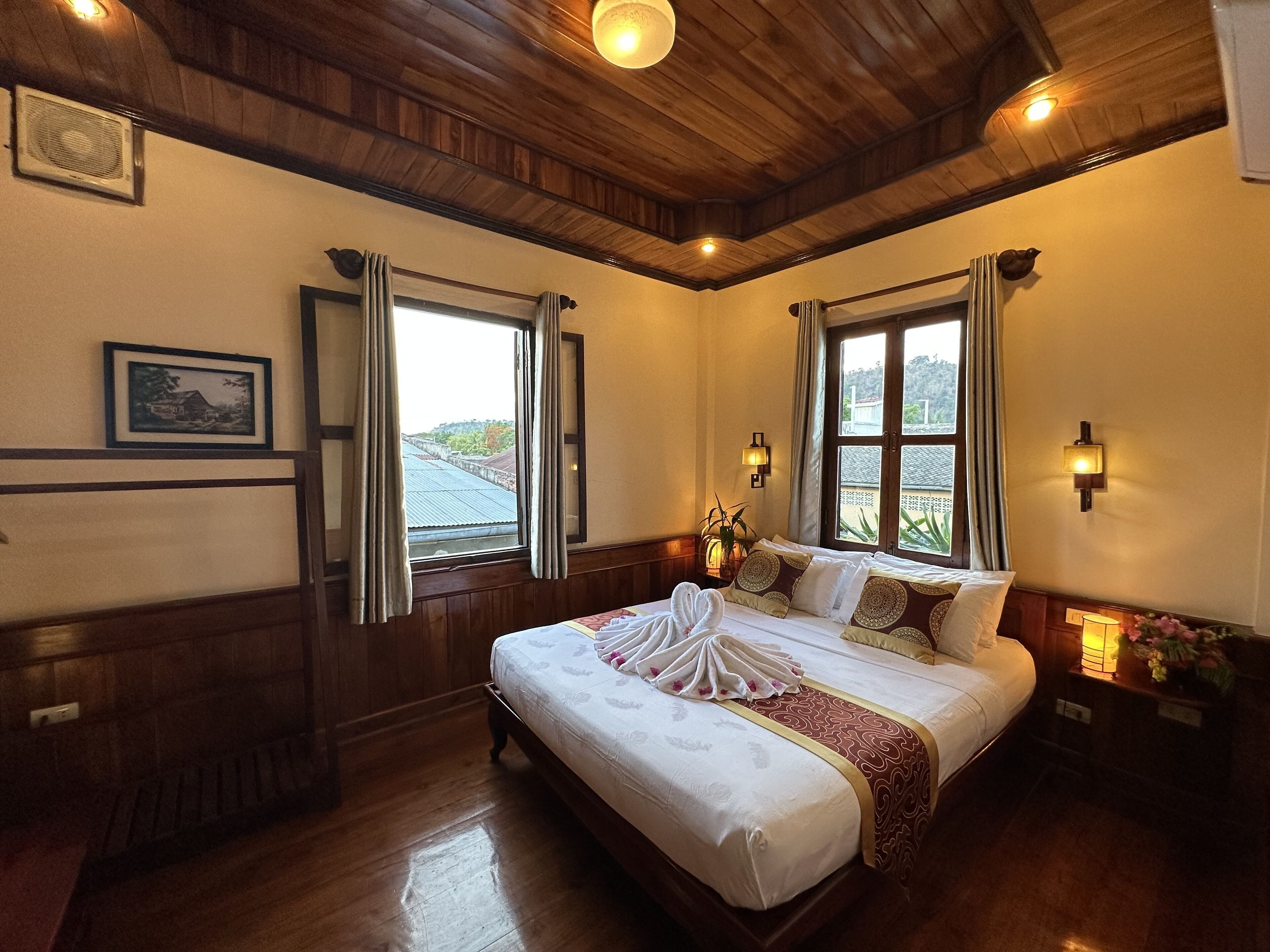 Habitación de hotel con una cama grande, techo de madera, dos ventanas con cortinas, una mesita de noche con lámpara y una lámpara de pared.