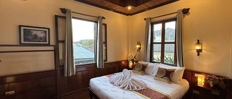 Habitación de hotel con una cama grande, techo de madera, dos ventanas con cortinas, una mesita de noche con lámpara y una lámpara de pared.