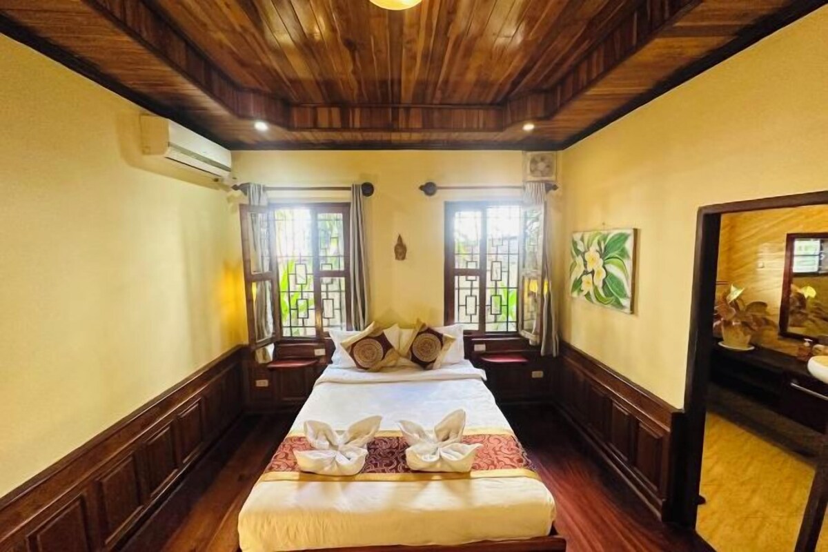 A hotel room with a large bed, two windows with curtains, a wooden ceiling, and a wall-mounted air conditioner.