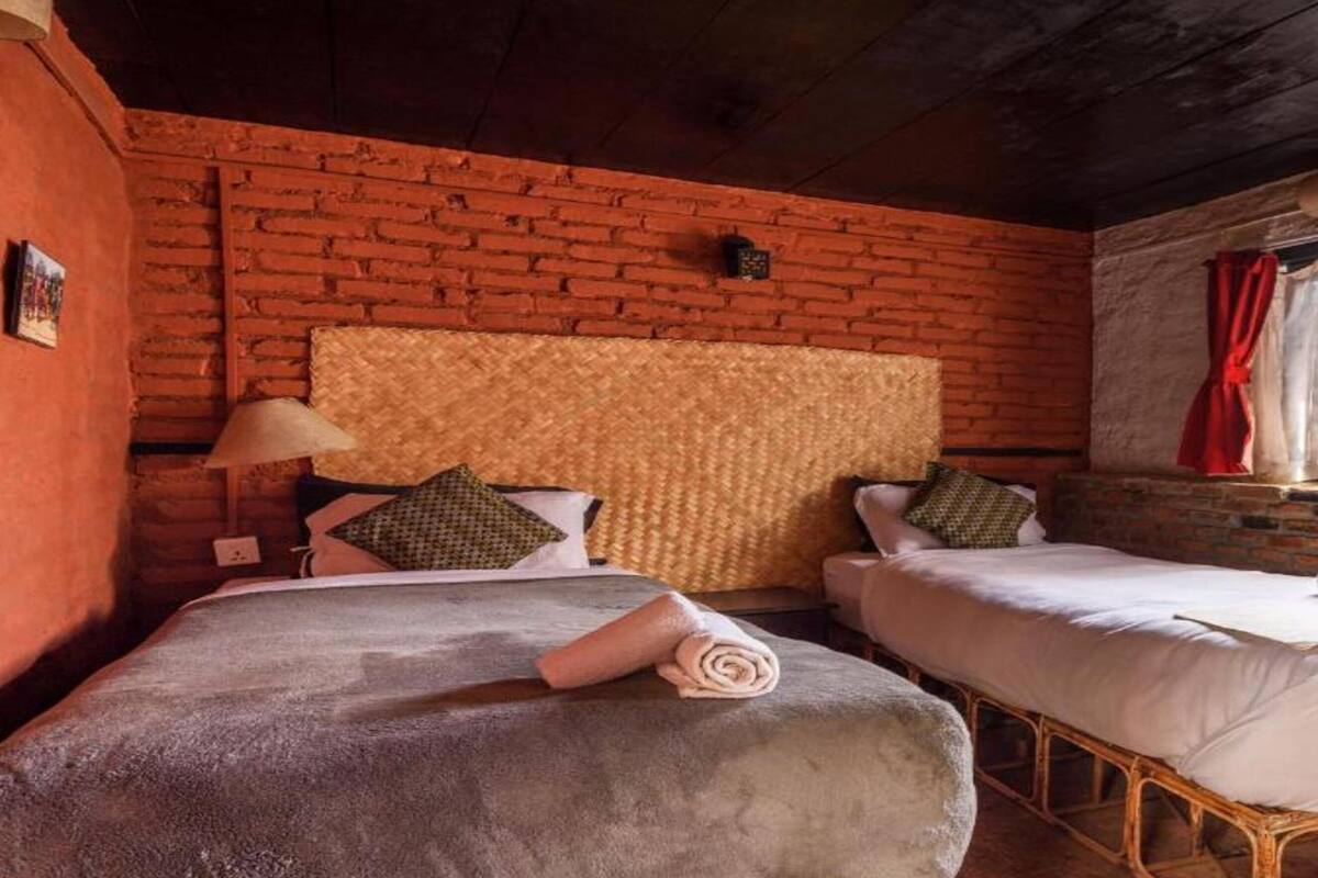 A room with two beds, a brick wall, a window with red curtains, and a woven headboard.