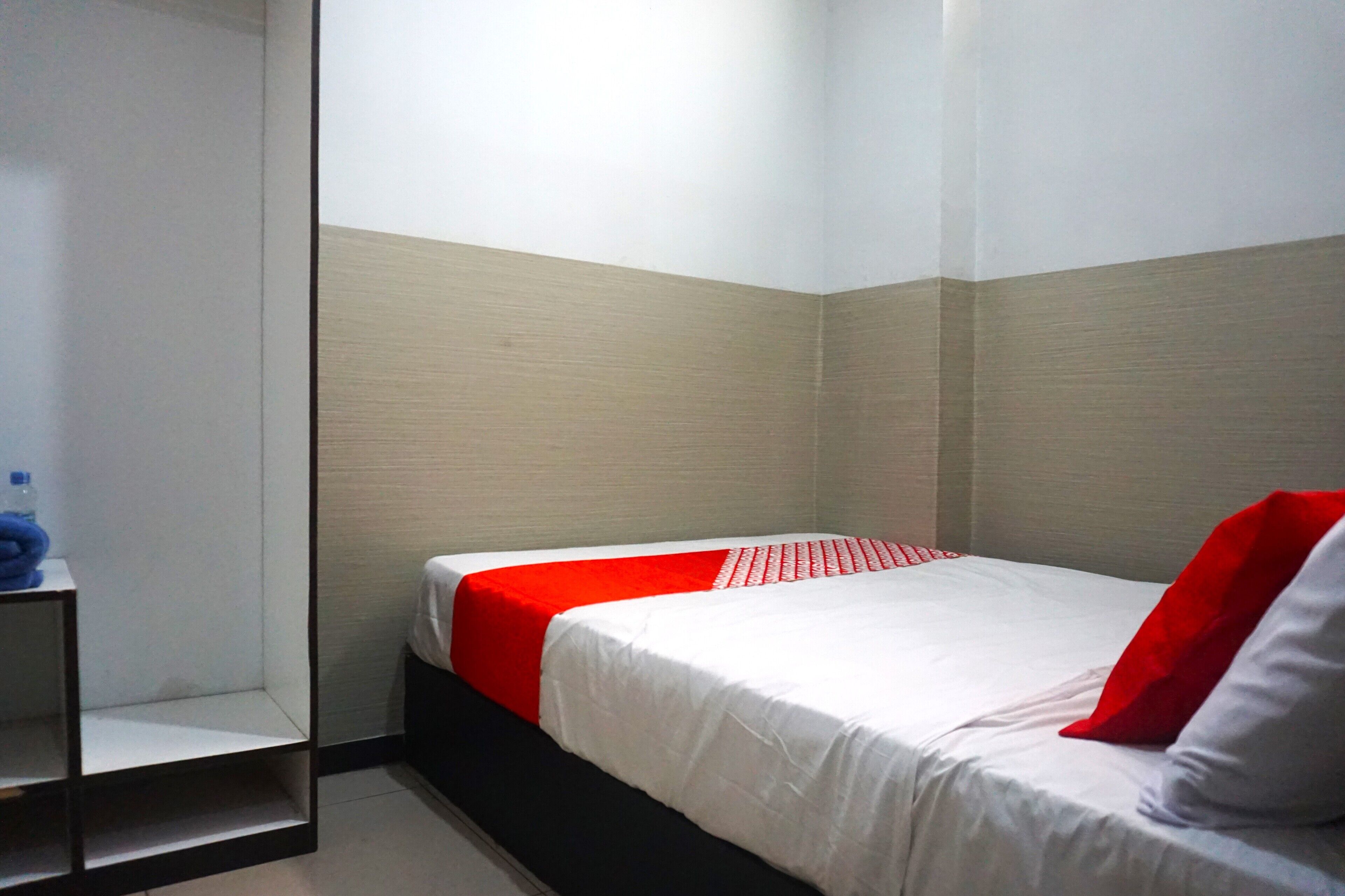 A hotel room with a bed, a red and white bedspread, a black shelf, and a white wall.