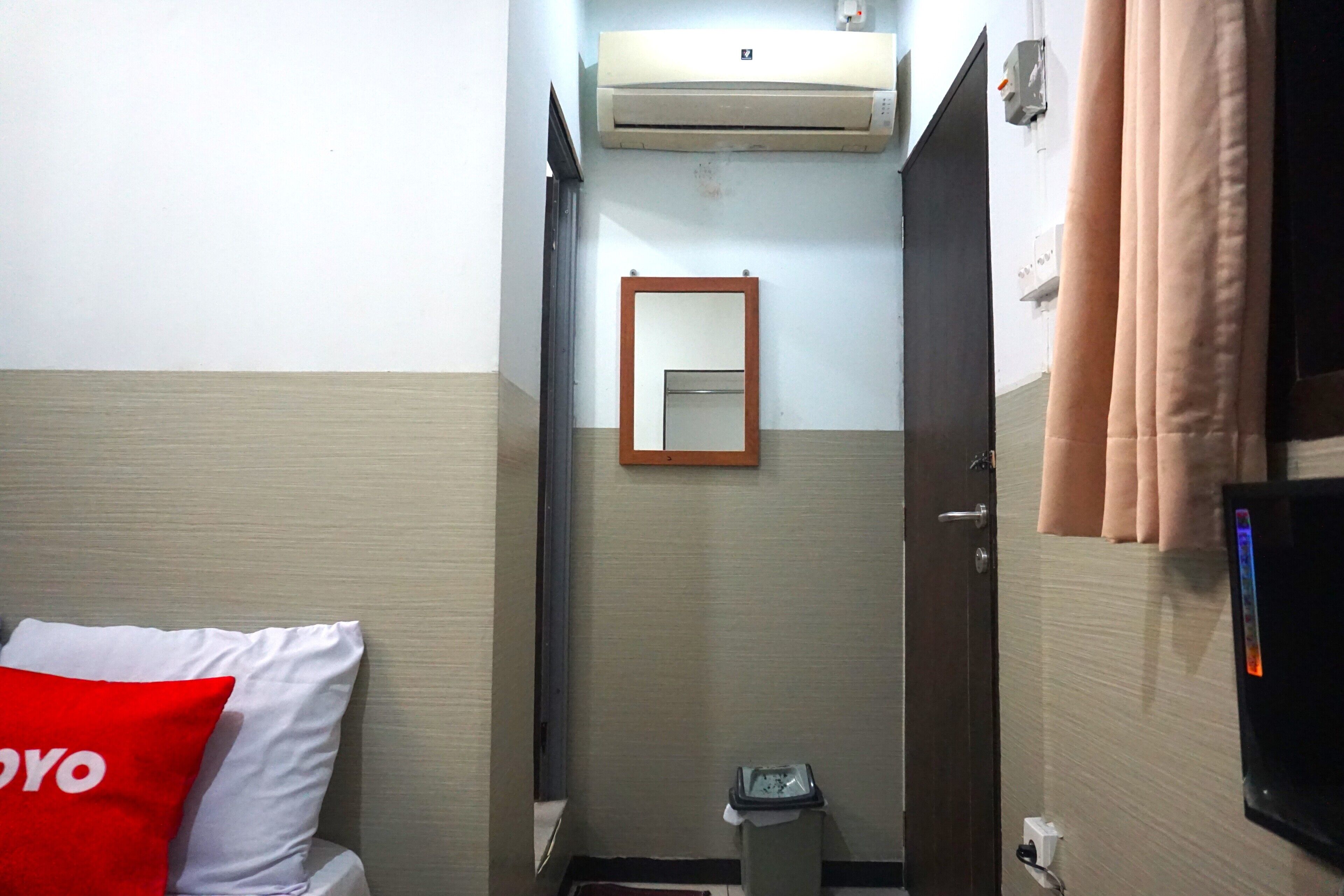 A small hotel room with a bed, a red pillow, a wall-mounted air conditioner, a mirror, a small trash bin, and a TV on the wall.
