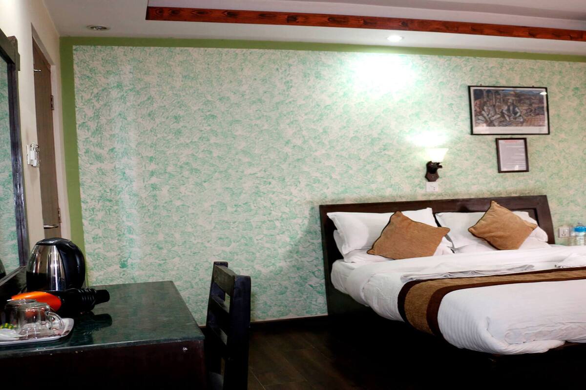 A hotel room with a bed, a desk with a kettle, a chair, and a wall-mounted picture.