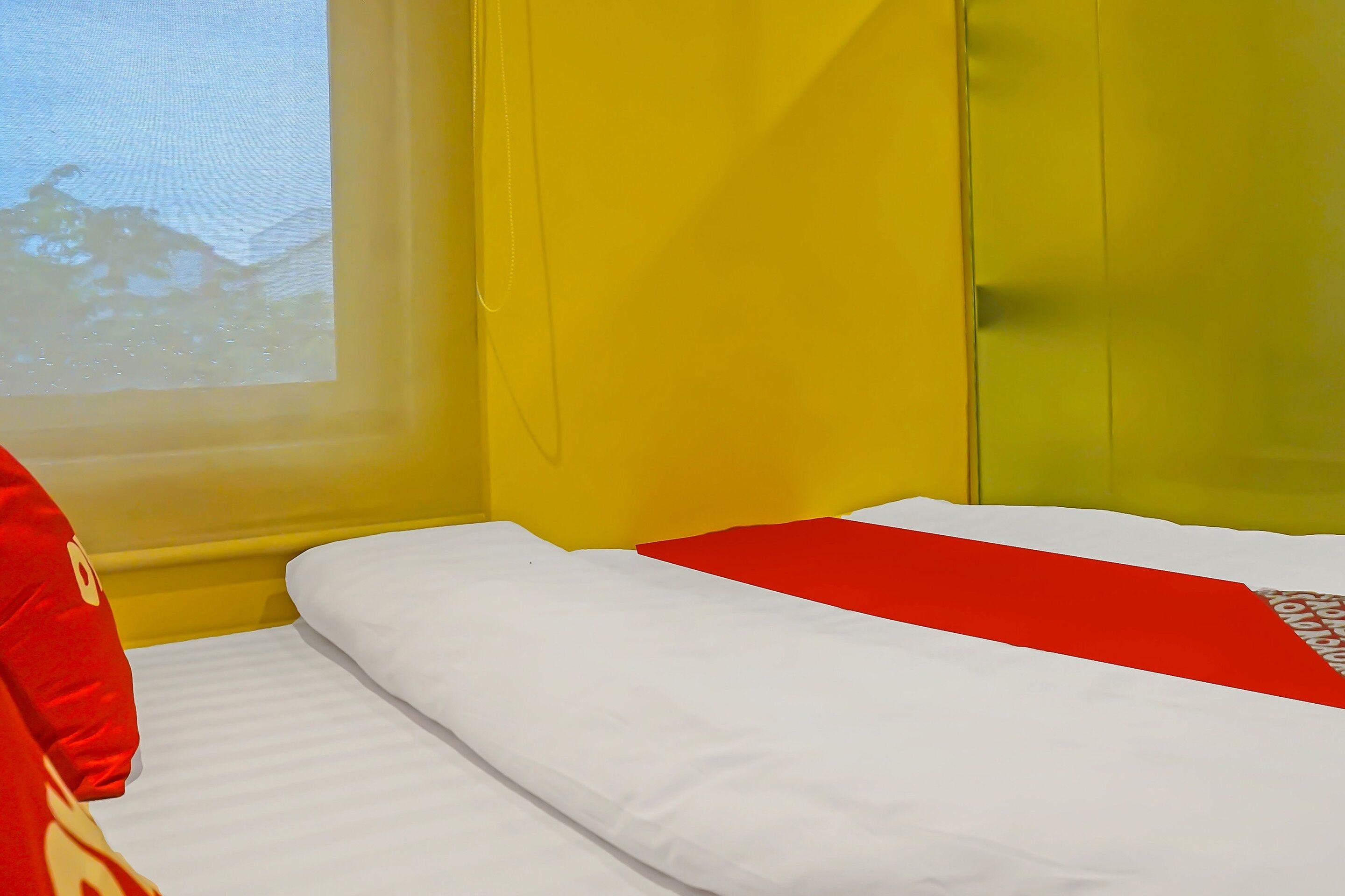 Una cama con ropa de cama blanca y roja, una pared amarilla y una ventana con vistas a árboles.