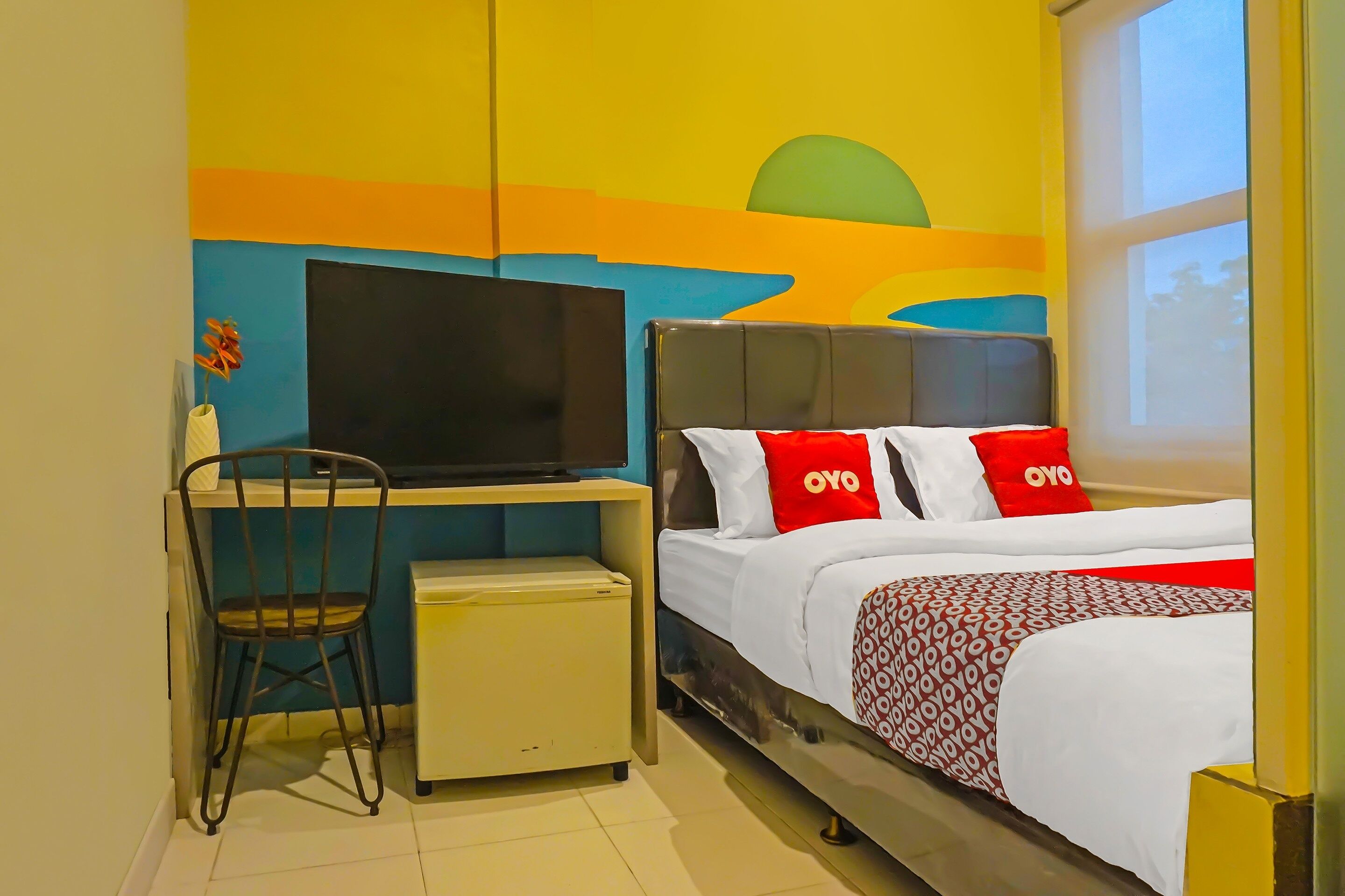 A hotel room with a bed, a desk with a chair, a television, and a wall with a colorful mural.