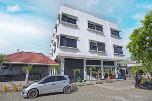 Un edificio de varios pisos con fachada blanca y marcos de ventana negros, un coche estacionado al frente y pequeñas palmeras alrededor de la propiedad.