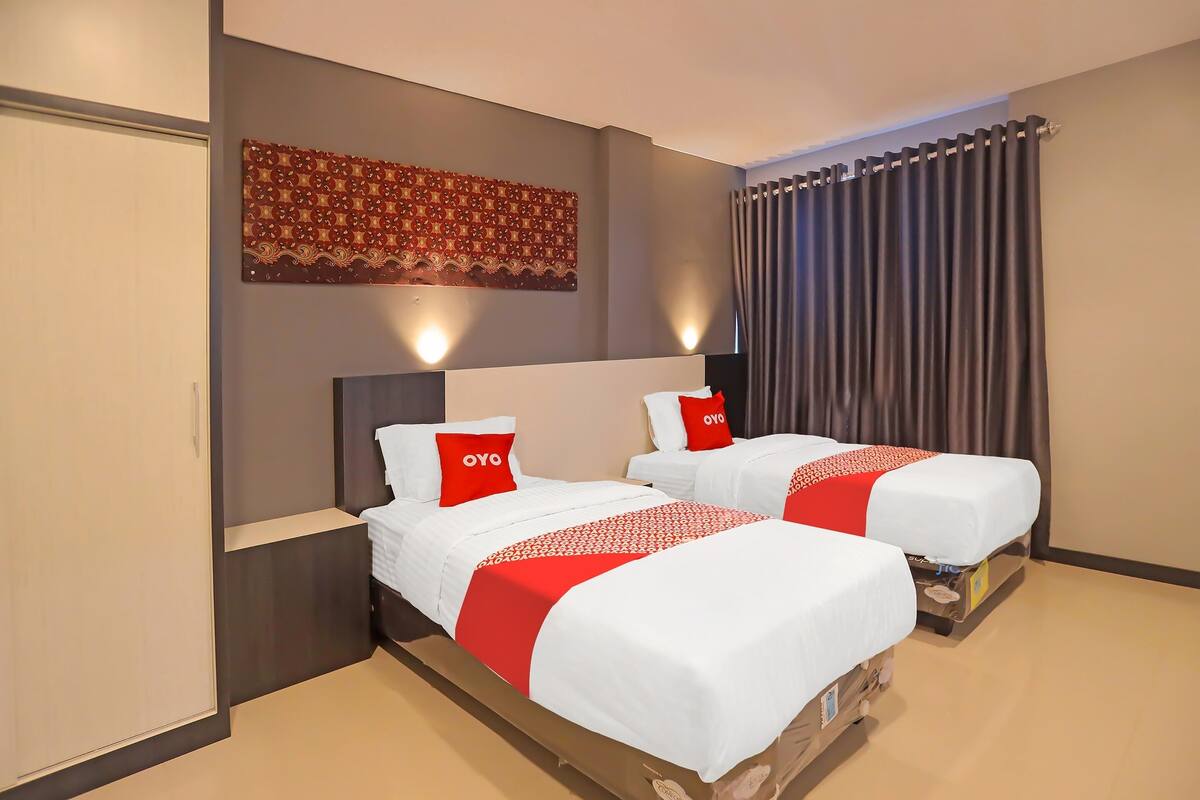 Quarto de hotel com duas camas, cada uma com roupa de cama branca e detalhes vermelhos, um grande painel decorativo na parede e uma cortina na janela.