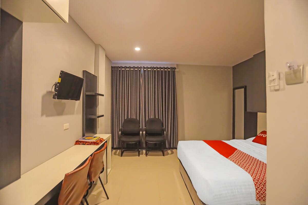 Quarto de hotel com uma cama, uma escrivaninha, duas cadeiras, uma televisão e uma cortina.