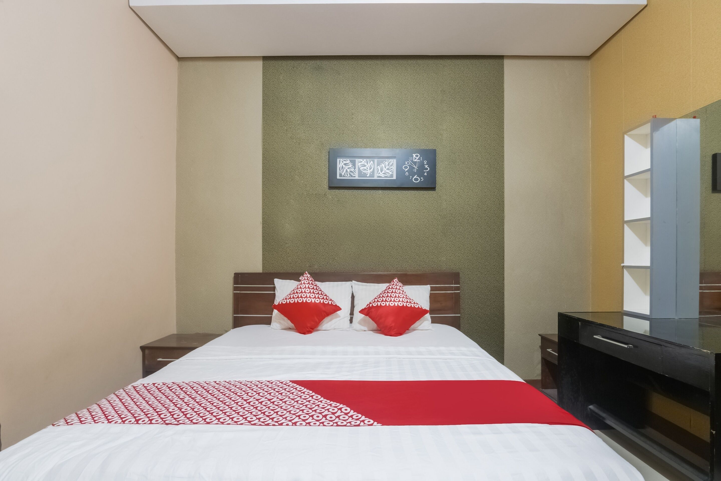 Una habitación de hotel con una cama, dos almohadas rojas, una mesita de noche, un televisor y un cuadro colgado en la pared.