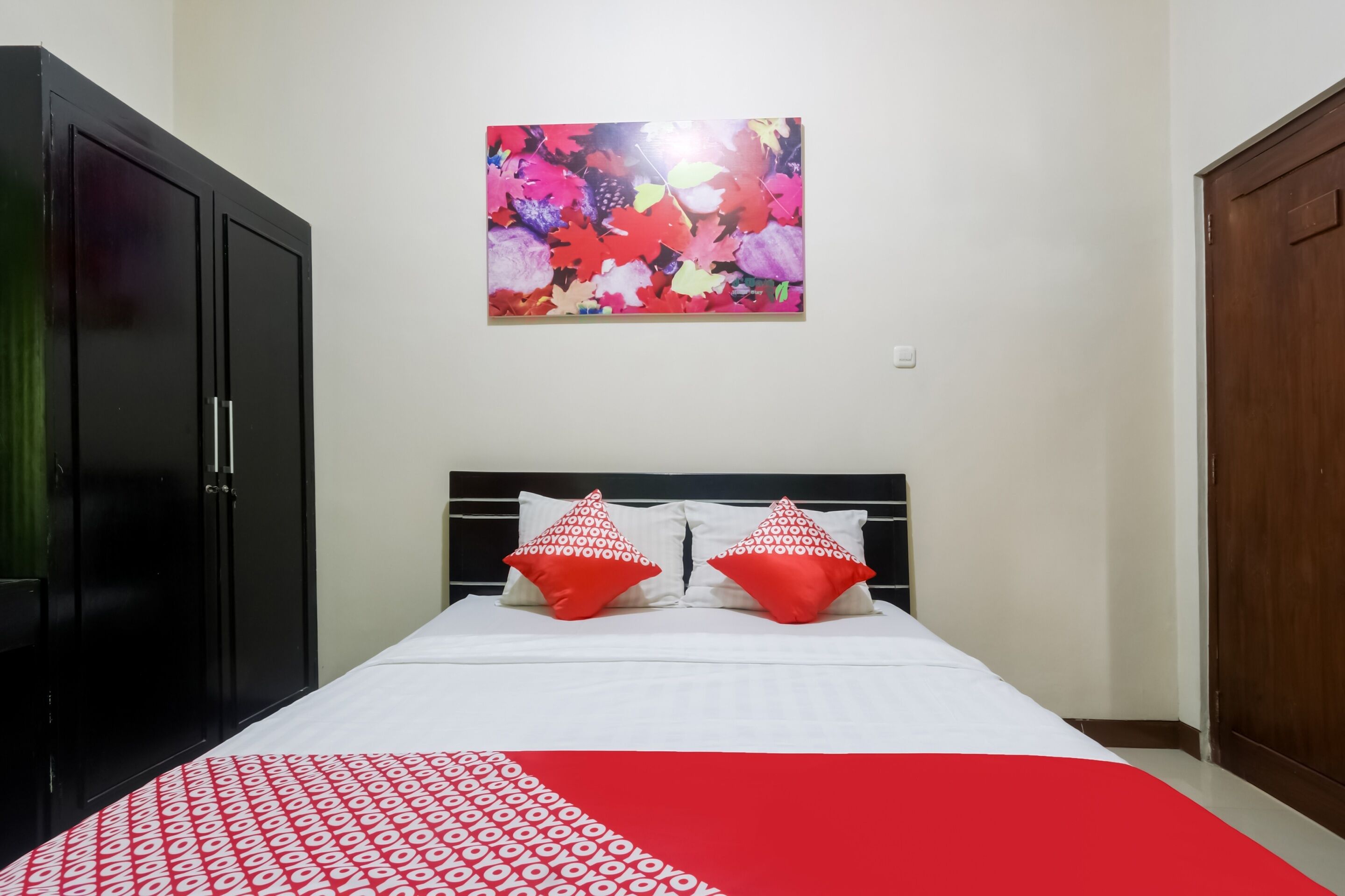 Un lit bien fait, recouvert d’une couvre-lit à motifs rouges et blancs, avec deux oreillers rouges ornés de motifs blancs, et une œuvre d’art florale aux couleurs vives accrochée au mur au-dessus du lit.