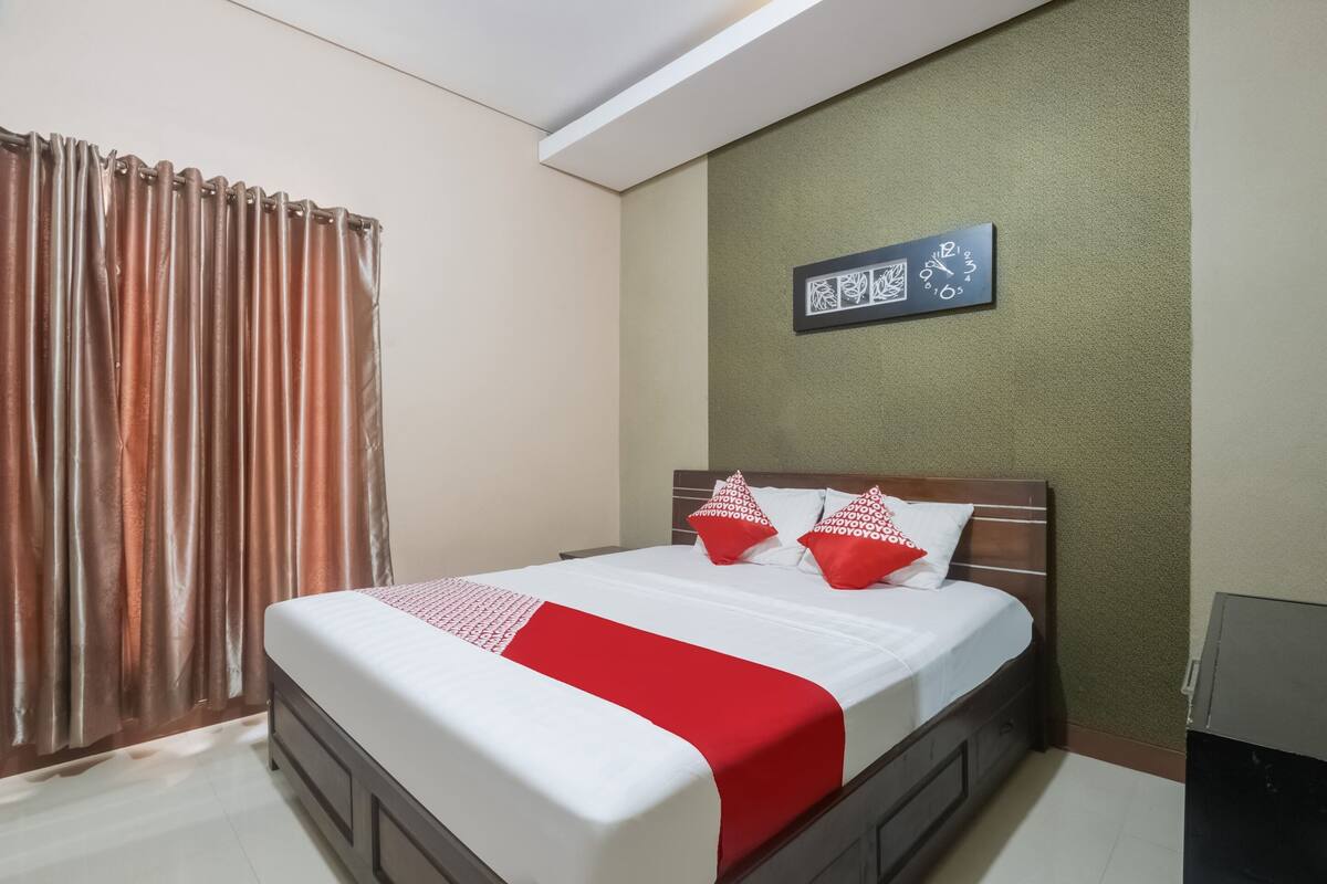 A neatly made bed with a red and white bedspread and pillows, a clock on the wall, and a window with curtains.