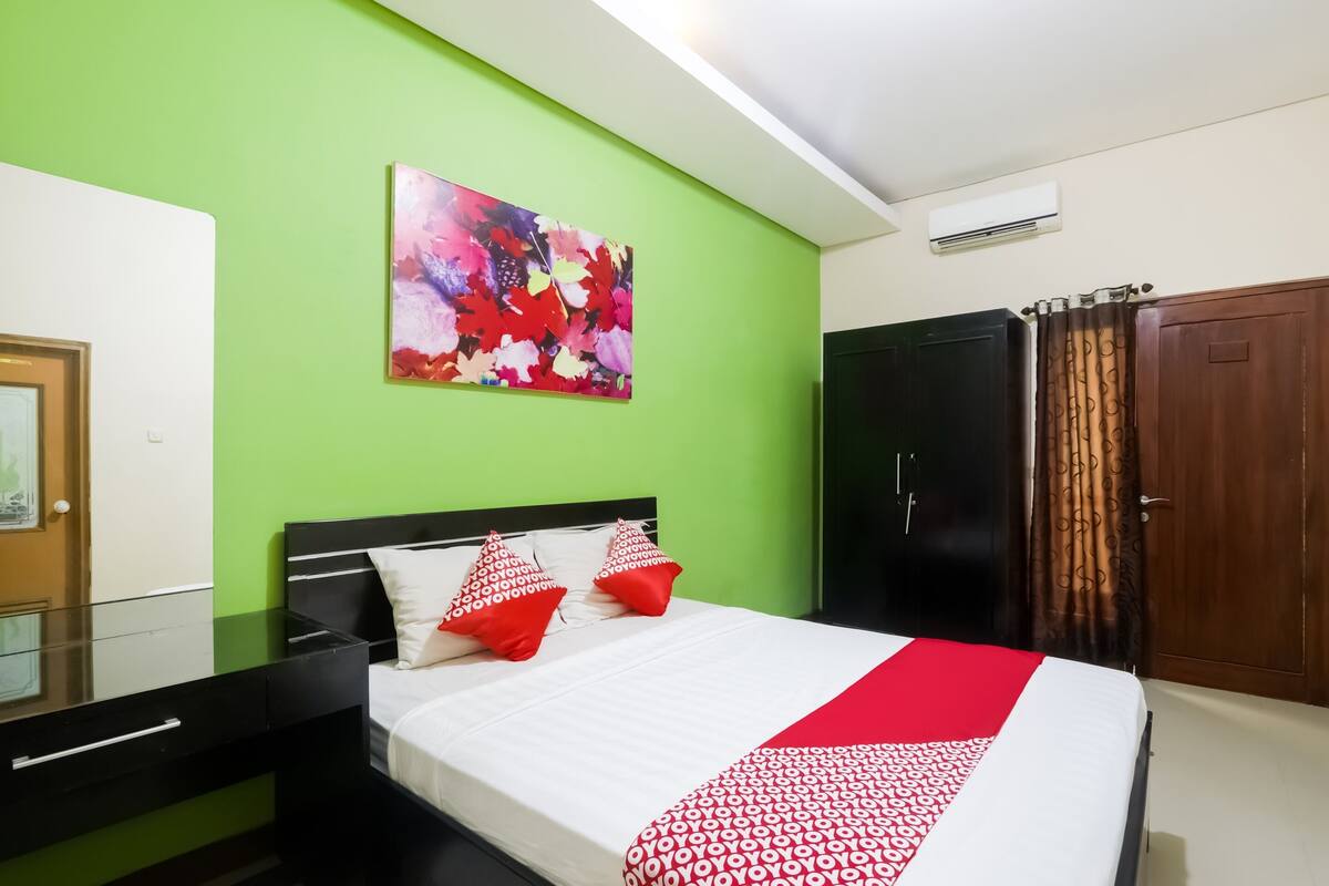 A hotel room with a bed, a green wall, a painting, a wardrobe, and a mirror.