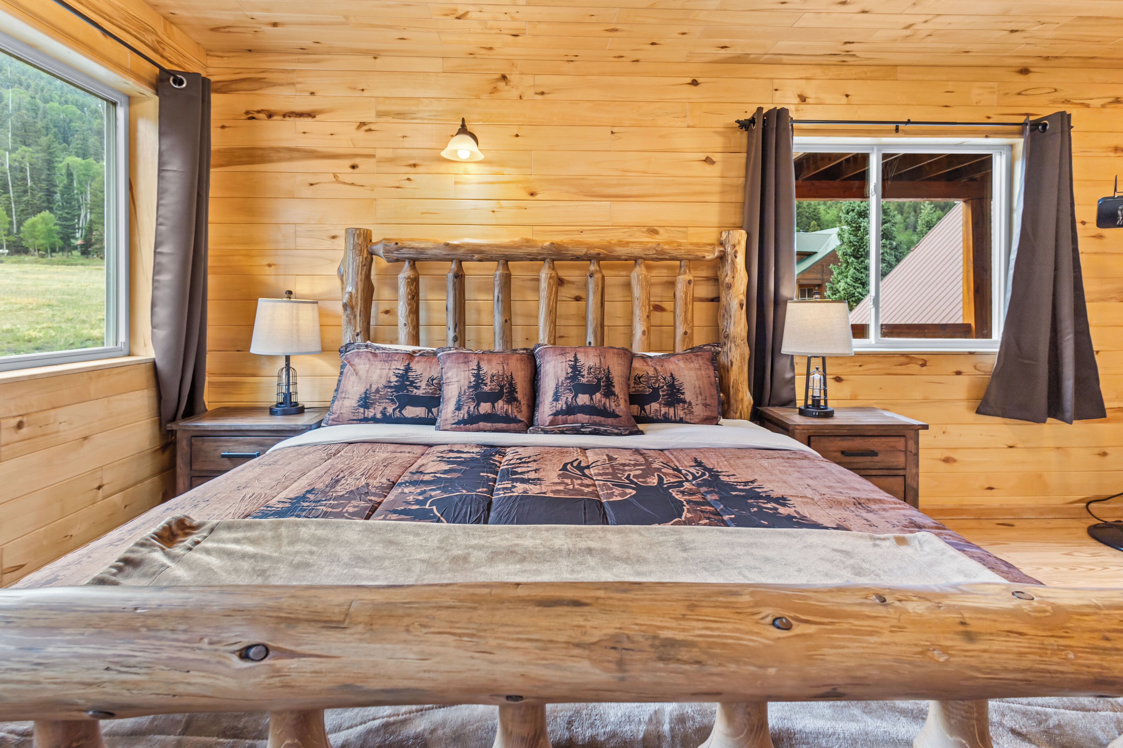 A wooden cabin bedroom with a bed, nightstands, a window with curtains, and a view of the outdoors.