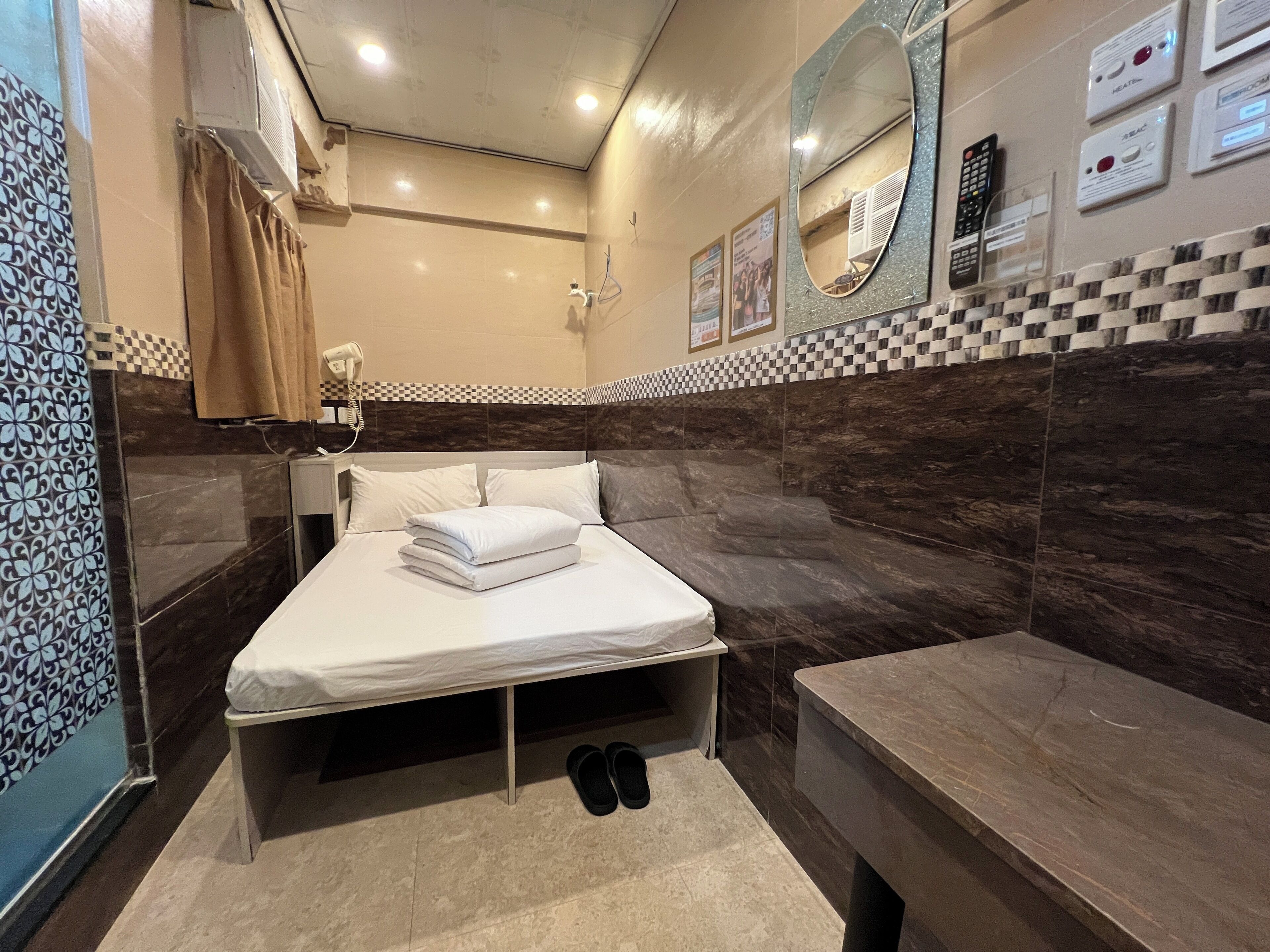 A hotel room with a bed, a bench, a mirror, and a wall-mounted control panel.