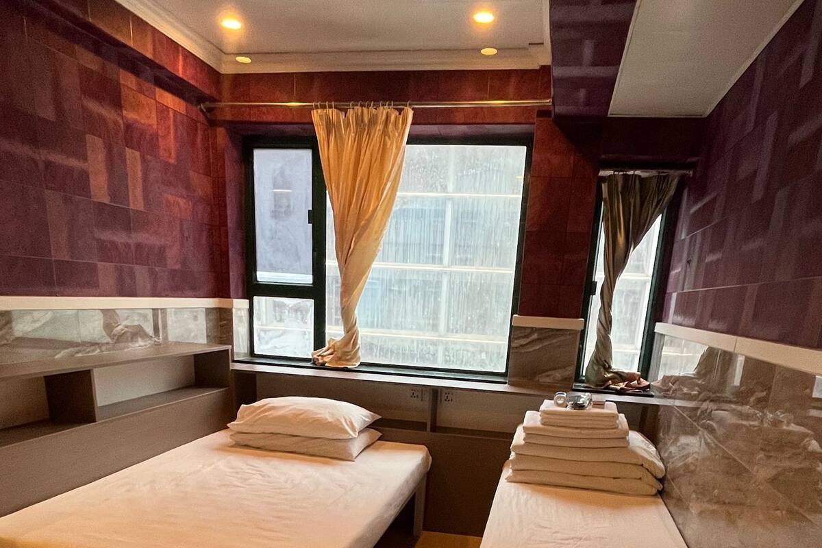 Une chambre d’hôtel avec deux lits, une fenêtre avec des rideaux, un plancher en bois et un petit tabouret.