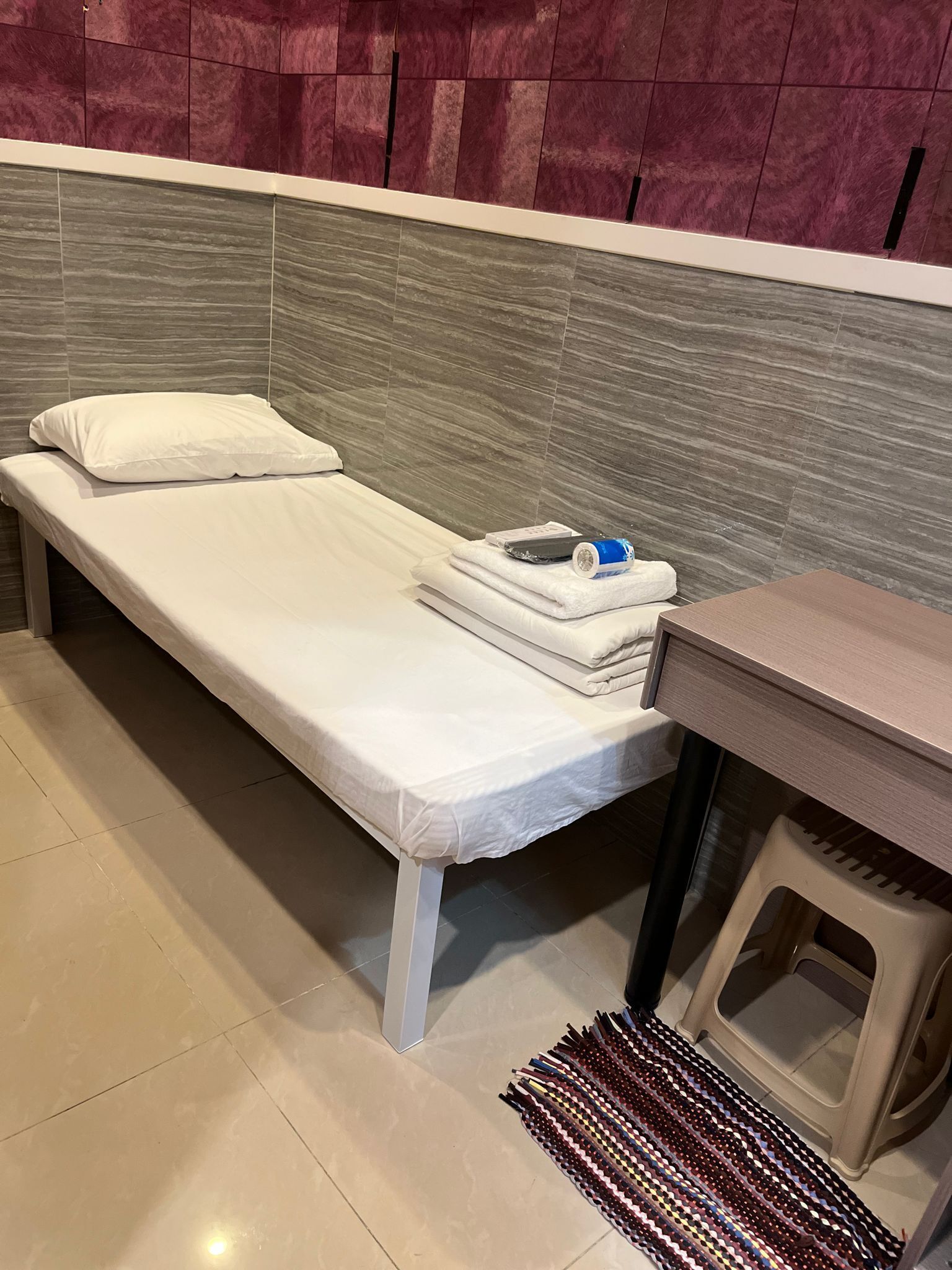 A small, single-bed room with a grey headboard, a white mattress, and a small bedside table with a towel and a remote control.