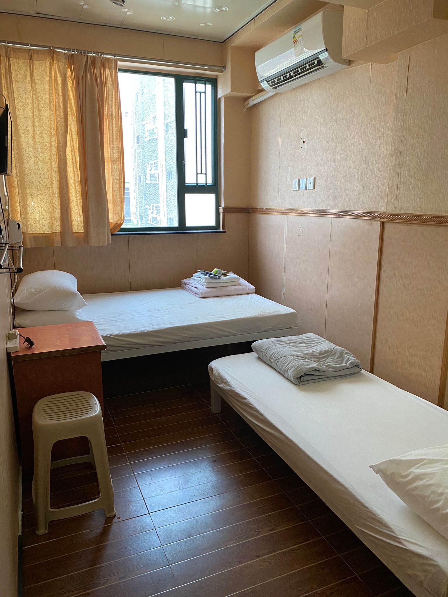 A small, compact room with a single bed, a small desk, a stool, and an air conditioning unit.