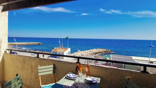Un balcon avec une table dressée pour le dîner, donnant sur une plage avec une digue rocheuse et un ciel bleu dégagé.