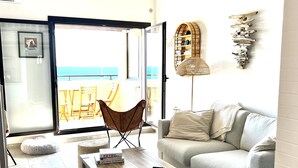 Un salon moderne avec un canapé gris, une table basse en bois et un fauteuil en osier. On peut admirer la mer à travers la porte vitrée.