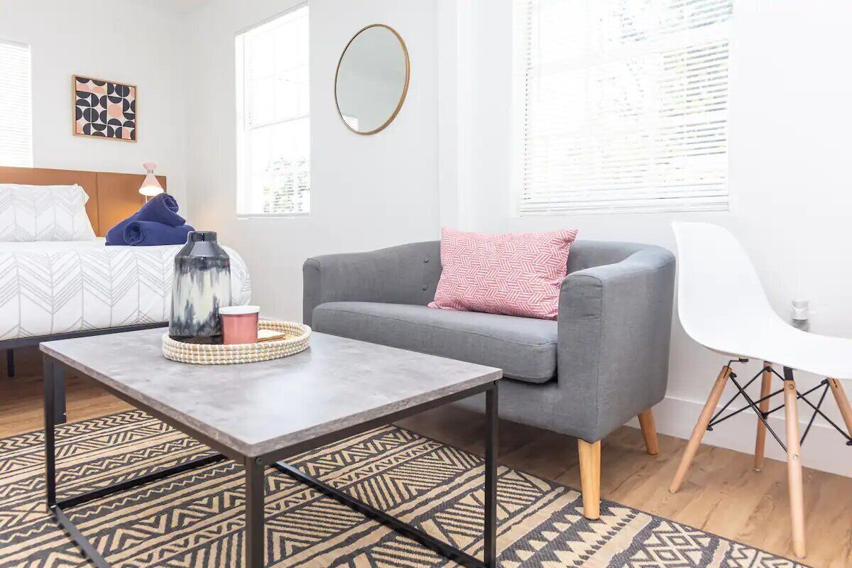 Quarto moderno com uma cama, sofá cinza, cadeira branca e uma pequena mesa com um objeto decorativo.