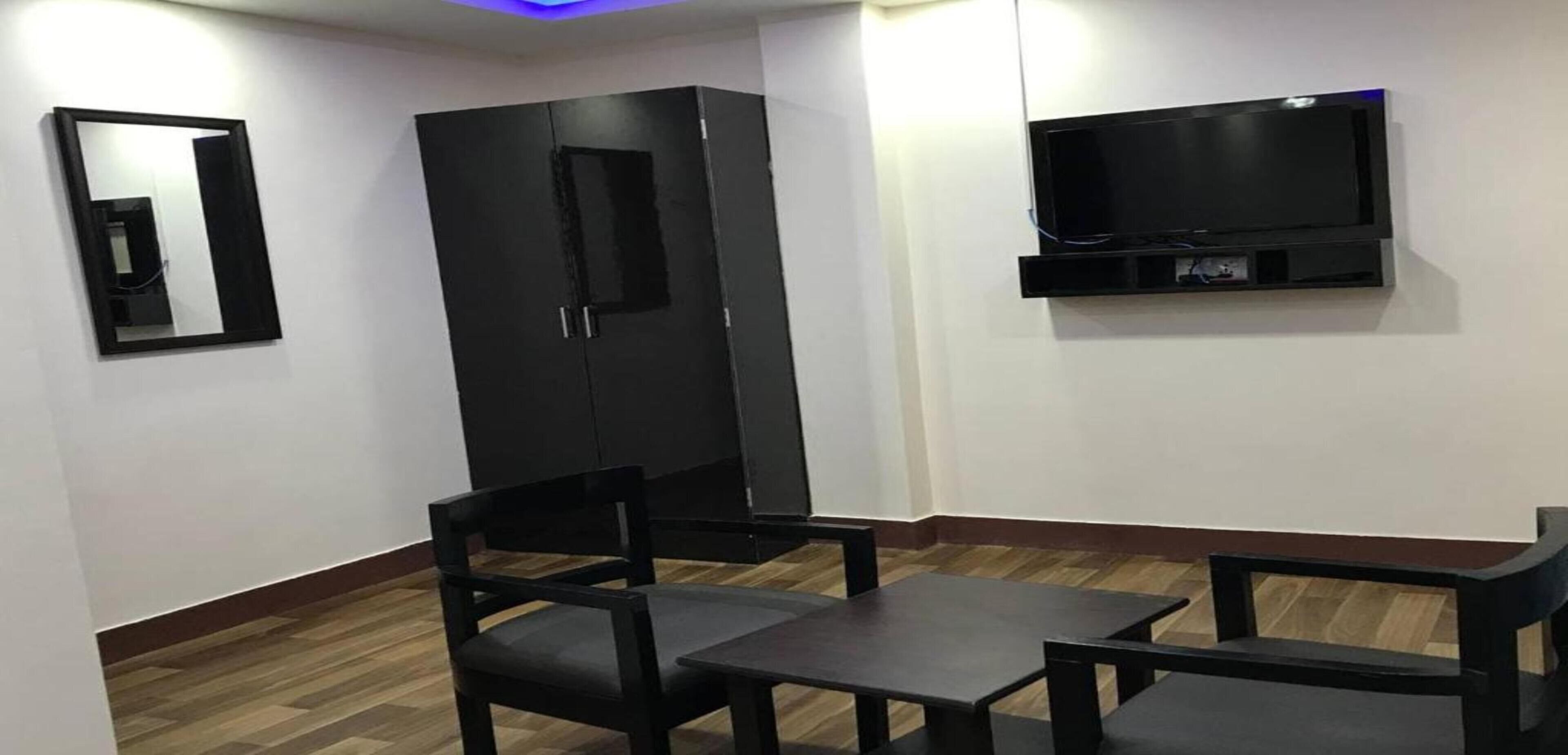 A room with a wooden floor, a black wardrobe, a black chair, a small table, and a mounted TV.