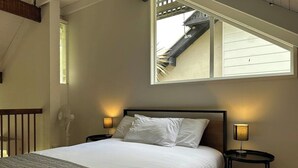 Ein ordentlich bezogenes Bett mit weißen Leinen, zwei Nachttischlampen, ein Holz-Kopfstück und ein Fenster mit Blick nach außen.