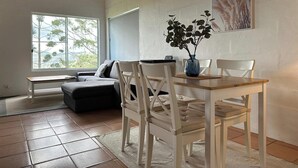 Ein Wohnzimmer mit einem Holzesstisch, zwei weißen Stühlen, einem grauen Sofa und einem großen Fenster mit Blick auf Bäume.
