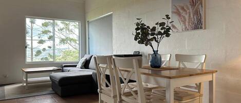 Un salon avec une table à manger en bois, deux chaises blanches, un canapé gris et une grande fenêtre donnant sur des arbres.