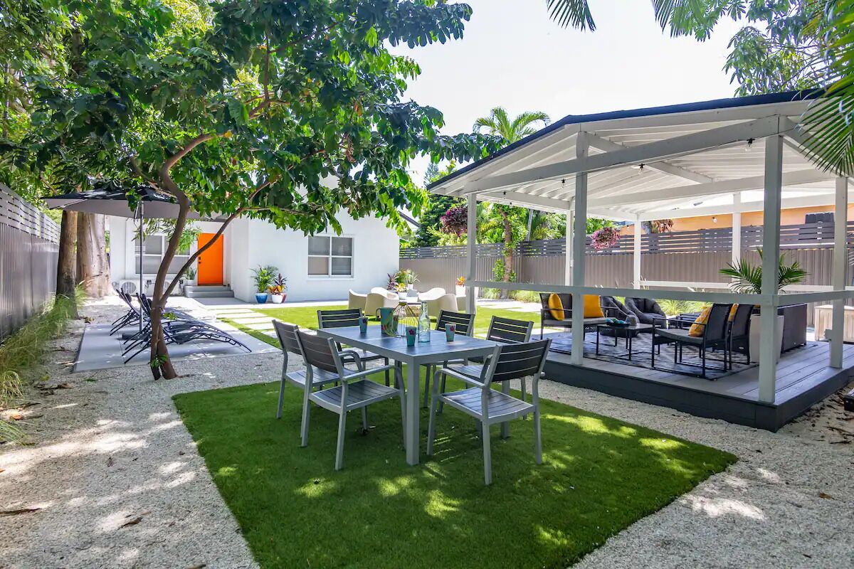 A covered patio area with a table and chairs, surrounded by a white fence and greenery.