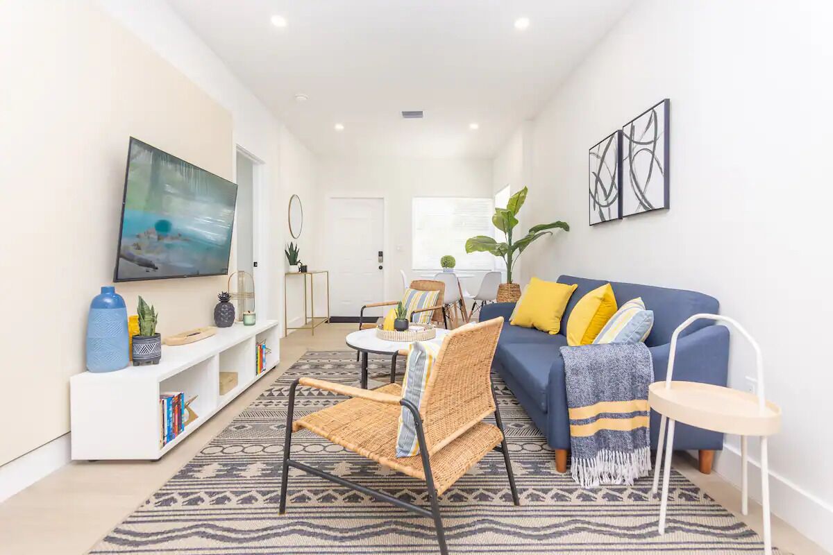 A modern living room with a blue sofa, a wicker chair, a white console table, and a TV mounted on the wall.