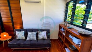 A room with a dark-colored sofa, a wooden side table, and a window with a view of greenery. - ZENHOUSE NIGHTCLIFF - 6BR Tropical Oasis (Nightcliff)