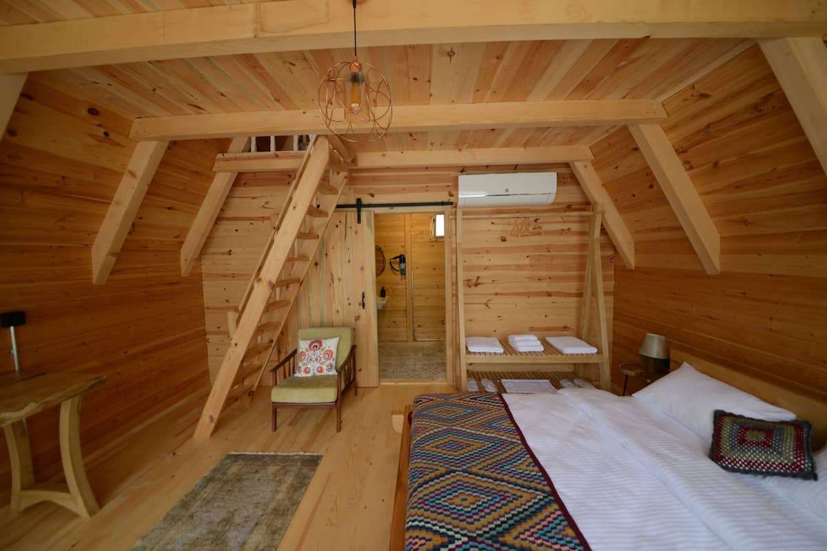 Un dormitorio de cabaña de madera con cama, una silla, una mesita, una escalera y un equipo de aire acondicionado.