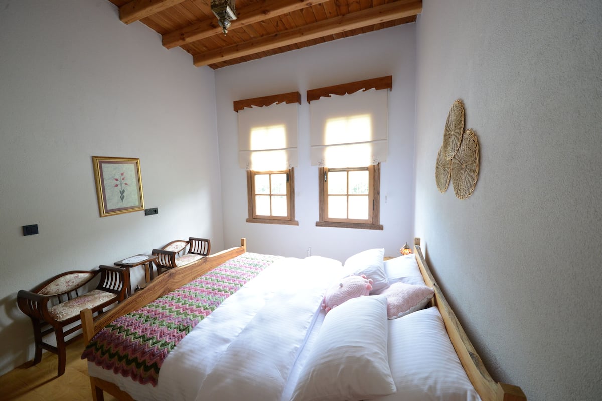 Un dormitorio con estructura de cama de madera, un banco de madera y dos ventanas con cortinas.